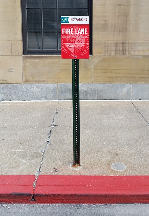 CSU Fire lane sign with water mark of white seal against a red background, somewhat transparent white seal. Small white text printed over the faded seal isn't readable from passenger side of car in lane.