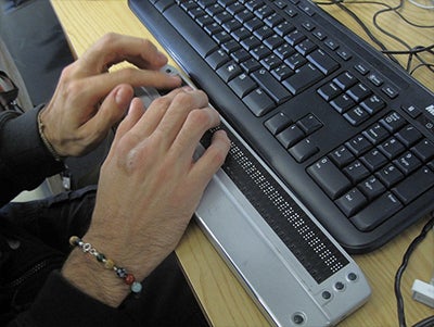 Hands on a refreshable braille display in front of a standard keyboard.