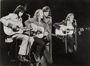 Four musicians, Crosby Stills Nash and Young, singing and playing guitars on a stage