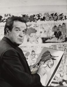 Cartoonist Al Capp sketching on a sketch pad. Cartoon strip wallpaper is on the wall behind him.