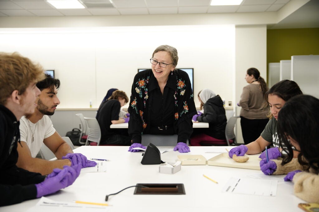 The photo captures a lively moment in a classroom where students are gathered around a central table, fully engaged in a hands-on learning activity with the curator, Sabine Kretzschmar, of the Education Art Collection of the Cleveland Museum of Art.