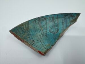 A turquoise marsh bowl fragment with dark-outlined floral motifs and red catalog markings.