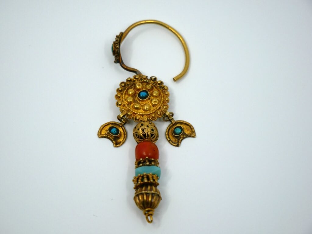A single ornate gold-toned earring with a circular top featuring small raised dots and a central turquoise stone. Two small crescent-shaped gold pieces with turquoise accents hang below, followed by a vertical stack of beads: a filigree gold bead, a red bead, a turquoise bead, and a larger ribbed gold bead at the bottom. The earring has a curved hook and an overall traditional, handcrafted look.