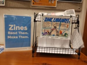 A zine display at a library sits on a wooden table. On the left, a blue sign reads, “Zines. Read Them. Make Them.” On the right, a black wire rack holds folded zines and informational handouts. A colorful poster on the front of the rack says “ZINE LIBRARY!” and explains that zines are self-published booklets that anyone can create about any topic. Craft supplies and zine-making imagery decorate the poster, and additional brochures are visible behind it.