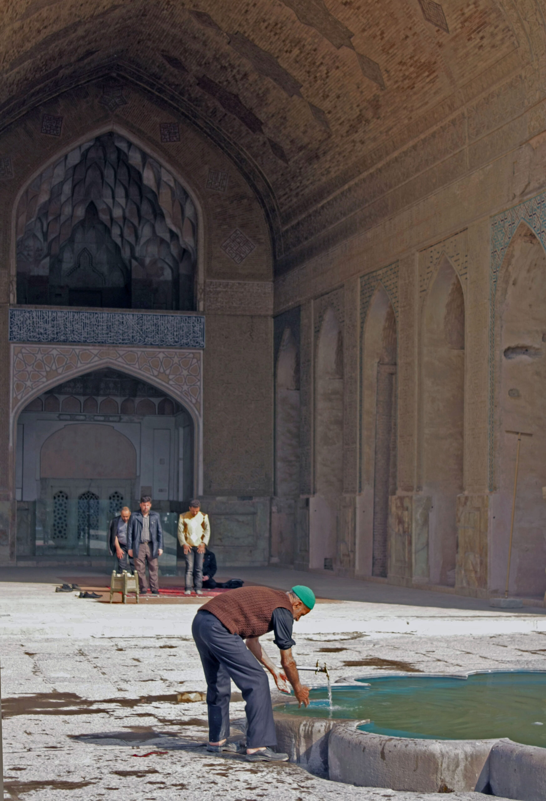Image of Man performing ablution, and men praying in the background, in the courtyard of a mosque in Iran.