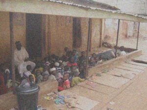 Islamic primary school students sitting on a porch with a teacher. Several foot tongs are placed near the entrance,