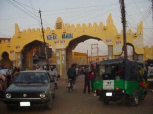 Yellow mud-brick gate, forty-feet high, wide enough to allow cars to pass through. The words "Kofar Wambai" are in the center of the gates. People and cars are in the foreground.