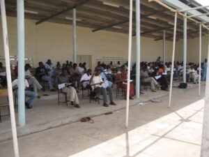 Dozens of students are seated and taking exams at desks in the shaded patio.