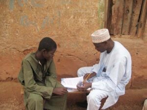 2 men seated on a bench outside, looking at papers.