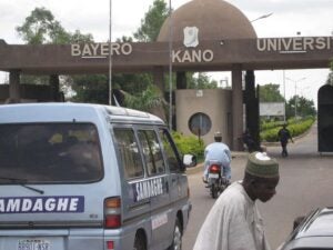 City entrance gate with the words: Bayero, Kano, and University on the header (background). In the forefront are a van, a man on a motorbike traveling in the direction of the gate. A man standing next to a car appears to be talking to the driver.