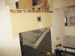 Words "Welcome to Department of Sociology" are printed on the wall overlooking the entrance to that area. A man is sitting on a bench near a staircase. A woman walks past the man on the bench.